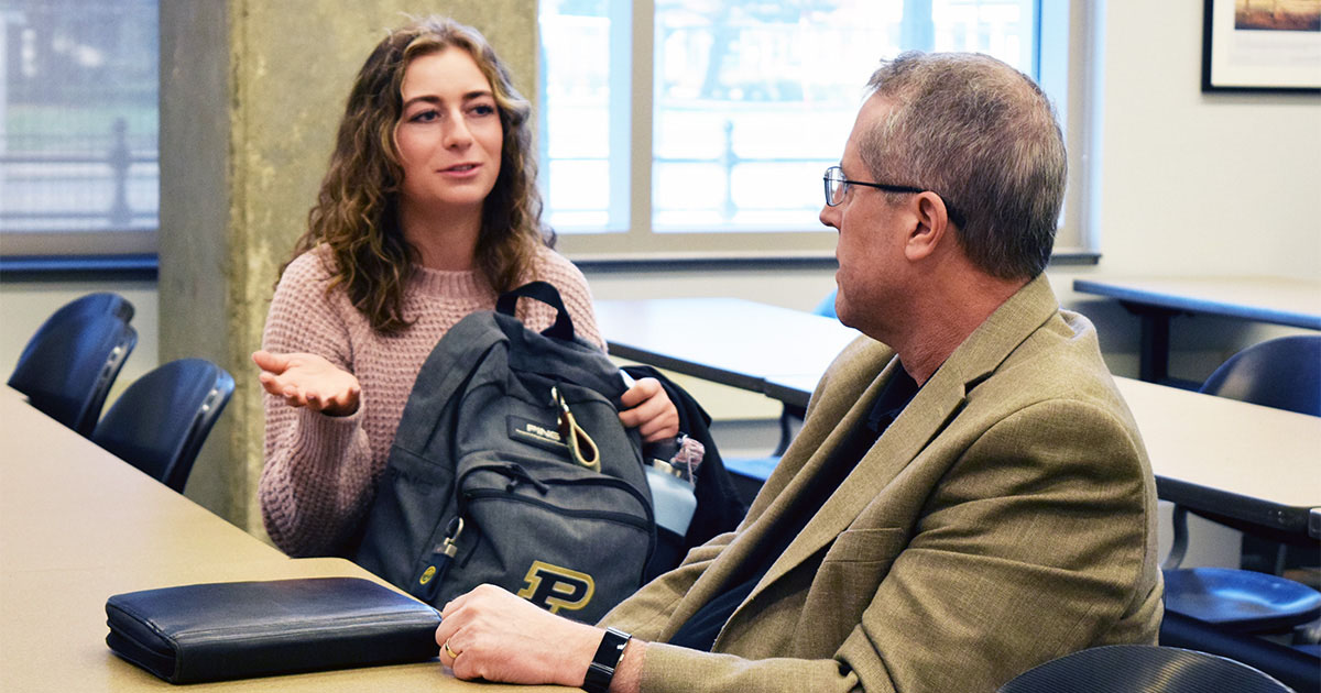 A female student seated at a table holding a backpack, talking to a middle-aged man in a suit seated next to her