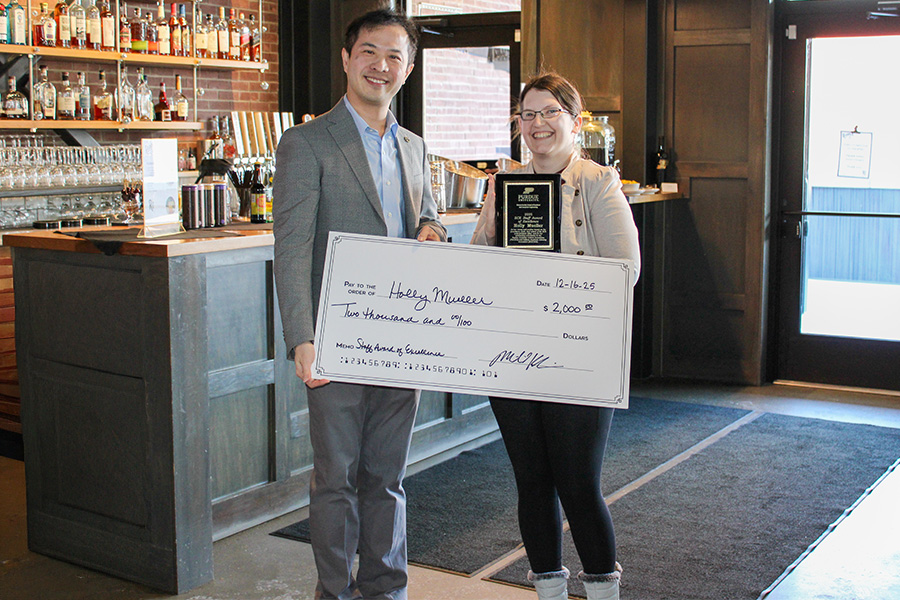 A smiling man and woman inside a bar, holding a large ceremonial check for $2,000 and a plaque. The interior is cozy, with shelves of bottles.