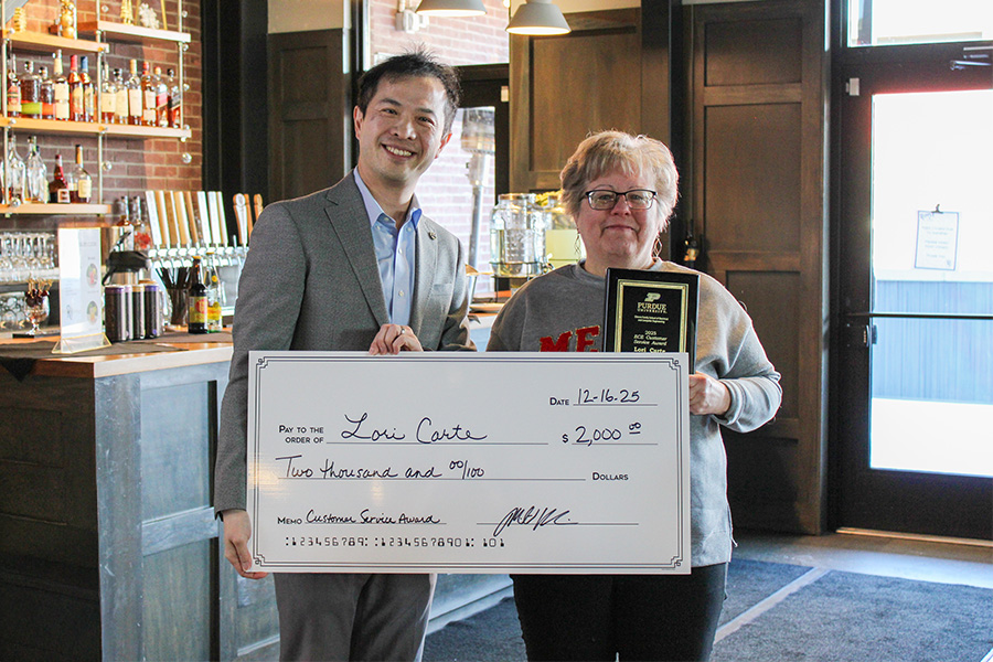 A smiling man in a suit stands beside a woman holding a large check for $2,000 and a plaque. They are in a warmly lit room with a bar in the background, conveying a celebratory and joyful atmosphere.